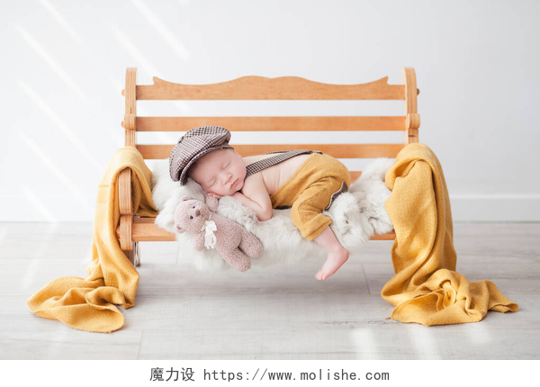 一个在照百天照的可爱宝宝一个穿着黄色裤子的新生婴儿和一个棕色流氓的帽子睡在一间明亮房间的木制长椅上，抱着一只玩具熊